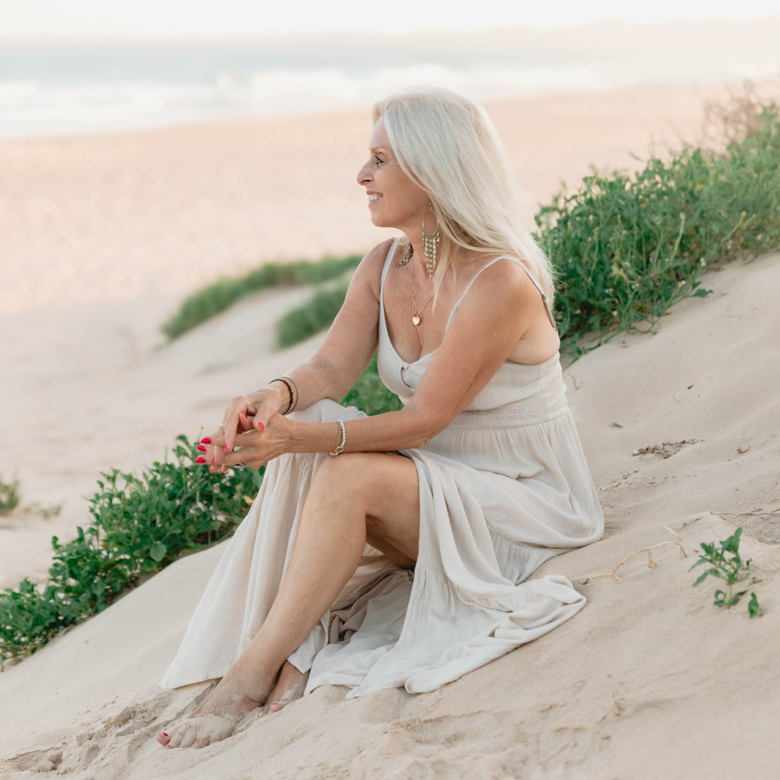 A serene scene of Val Hood, an Evidential Medium, sitting on the beach in a dress, embracing the peaceful coastal atmosphere.
